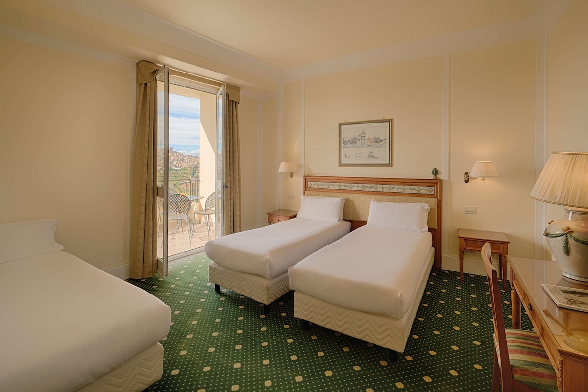 Een hotelkamer met twee enkele bedden, een groot raam met gordijnen, uitzicht op de stad, en een ingelijst schilderij aan de muur.