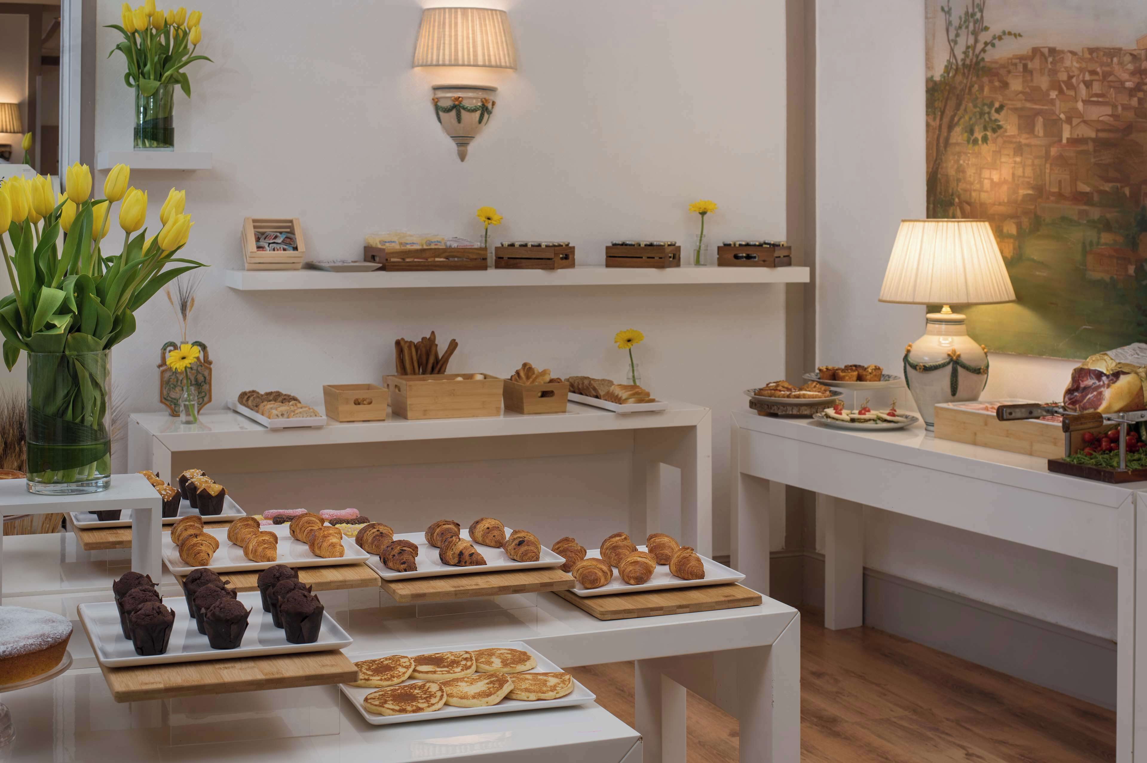 Montaje de bufé con diversos pasteles y postres sobre mesas blancas, rodeado de estantes en la pared con más alimentos y flores decorativas.