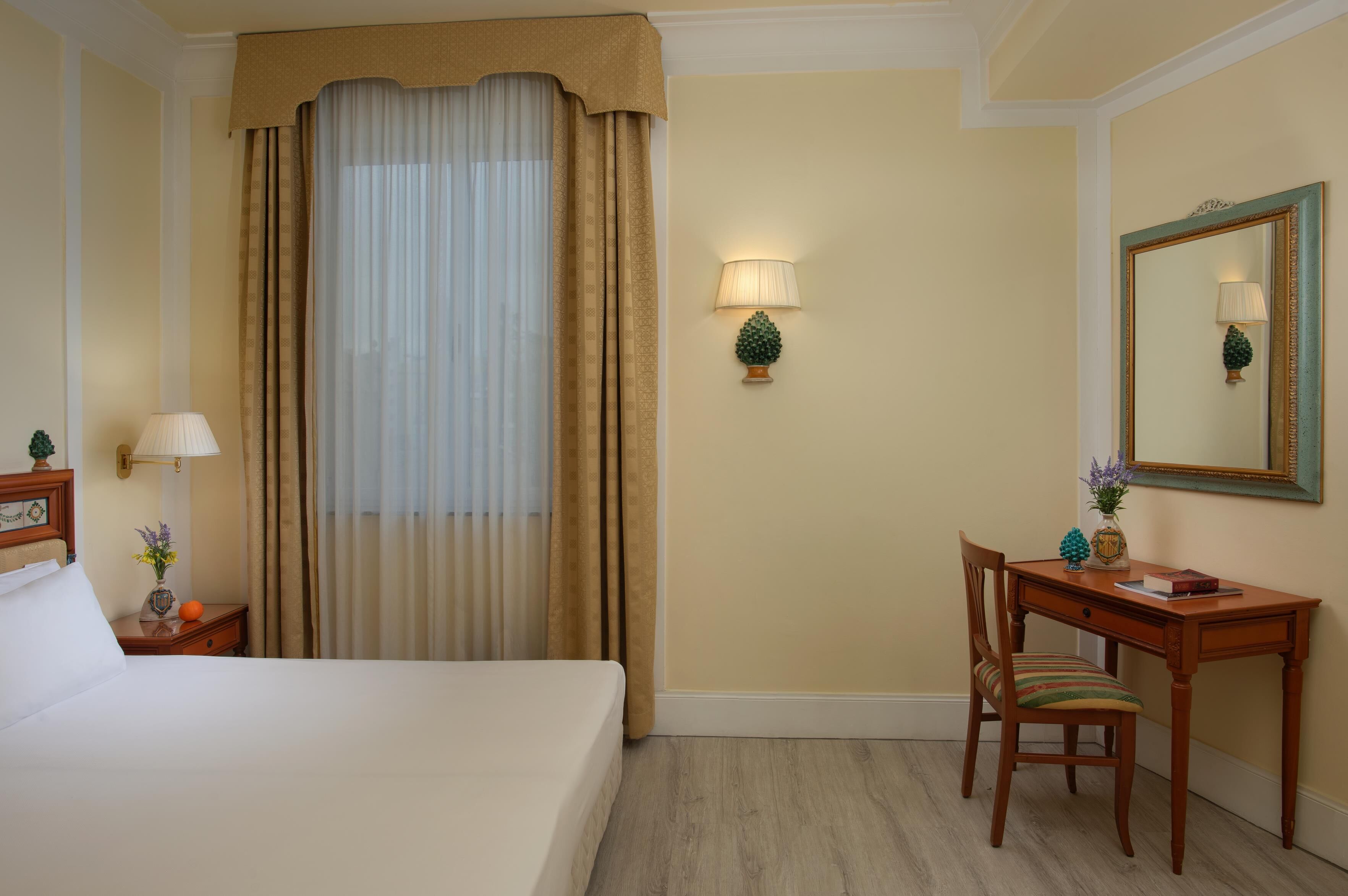 Una habitación de hotel con cama, escritorio con silla, lámpara y espejo.
