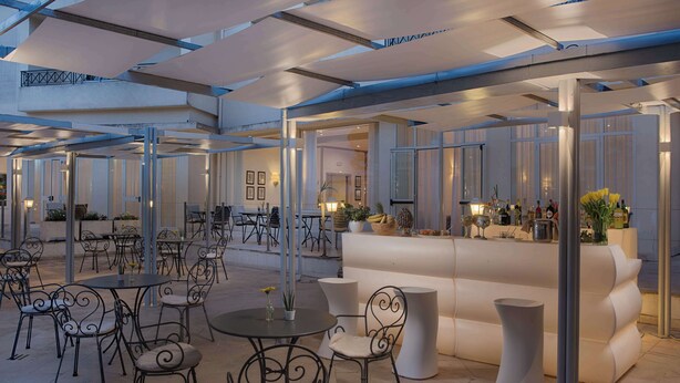 Ein moderner Außenbereich mit runden Tischen und Metallstühlen, eine Bar mit weißer Theke und ein großes weißes Sofa.
