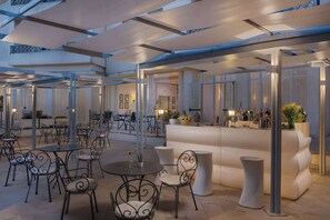Un espace extérieur moderne, aménagé avec des tables rondes et des chaises en métal, un bar doté d’un comptoir blanc et un grand canapé blanc.