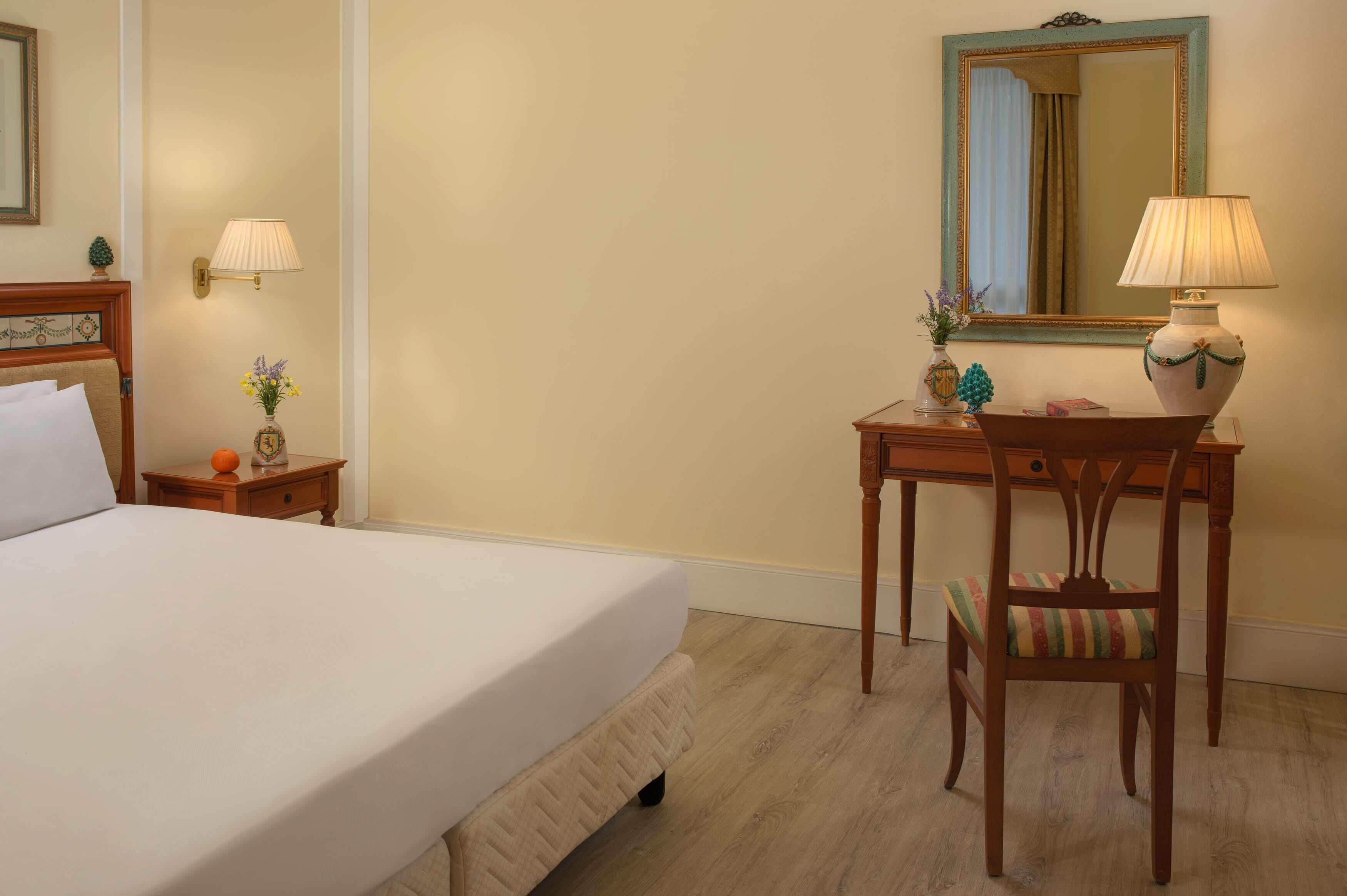 A hotel room with a bed, a desk, a chair, a lamp, a mirror, and a small vase with flowers.