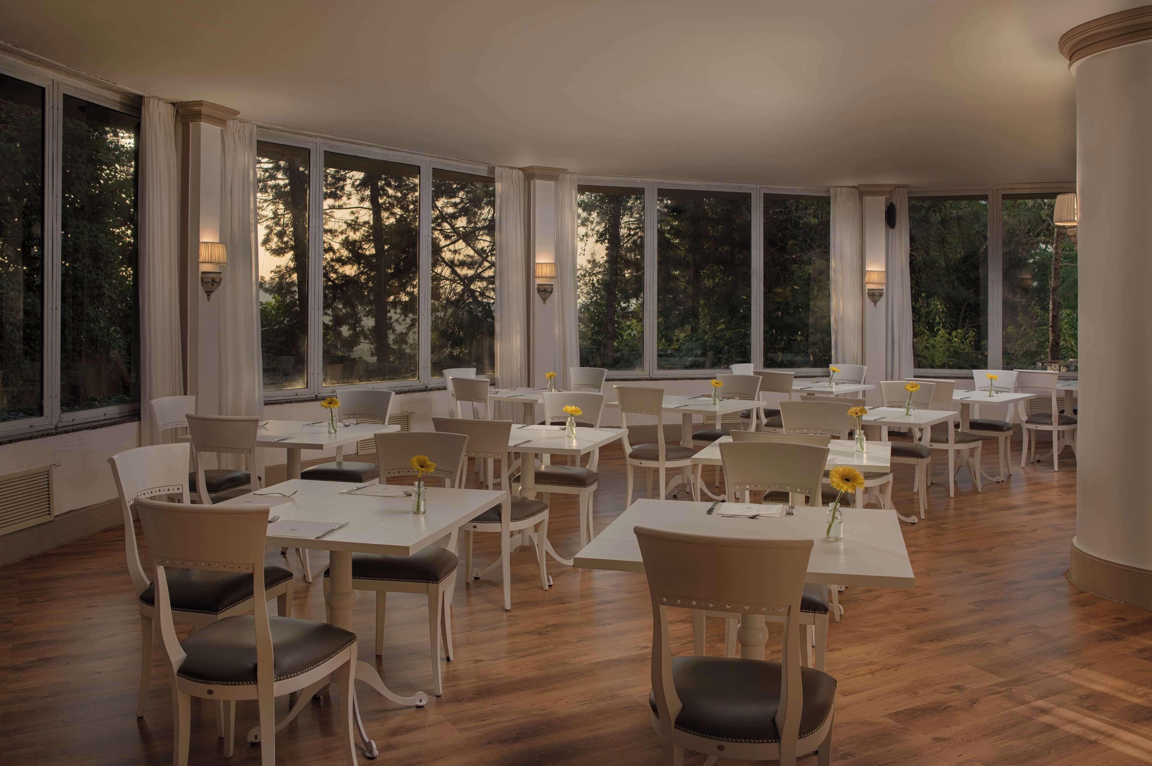 A spacious dining area with multiple tables and chairs, large windows with curtains, and wall-mounted lamps.