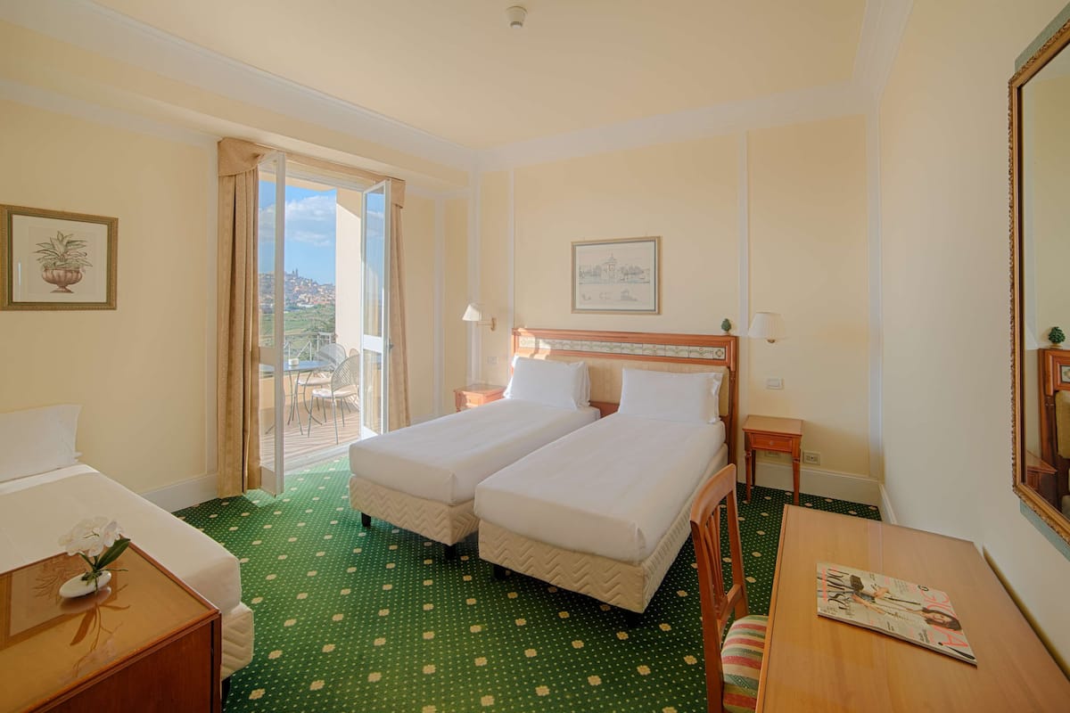 Een hotelkamer met twee enkele bedden, een houten nachtkastje, een kleintje tafeltje, een stoel, een balkon met uitzicht, en een fotokader aan de muur.