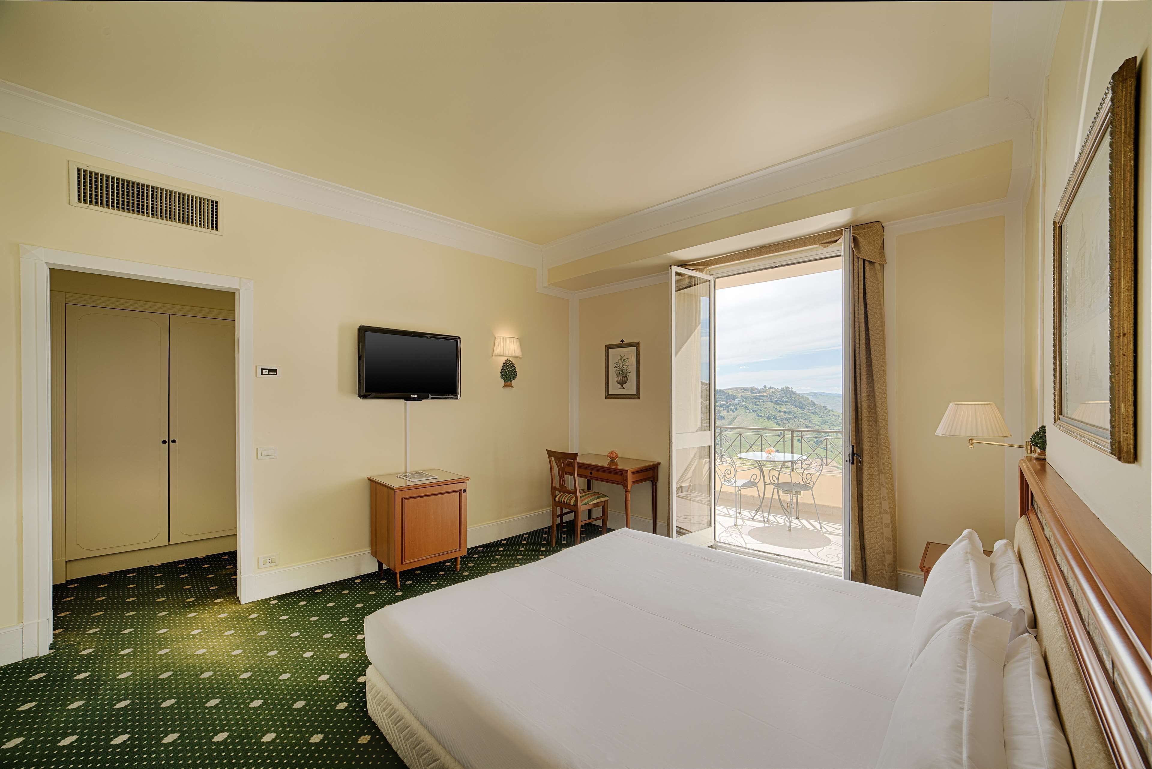 Habitación de hotel con cama, escritorio, silla, televisor y balcón con vistas.