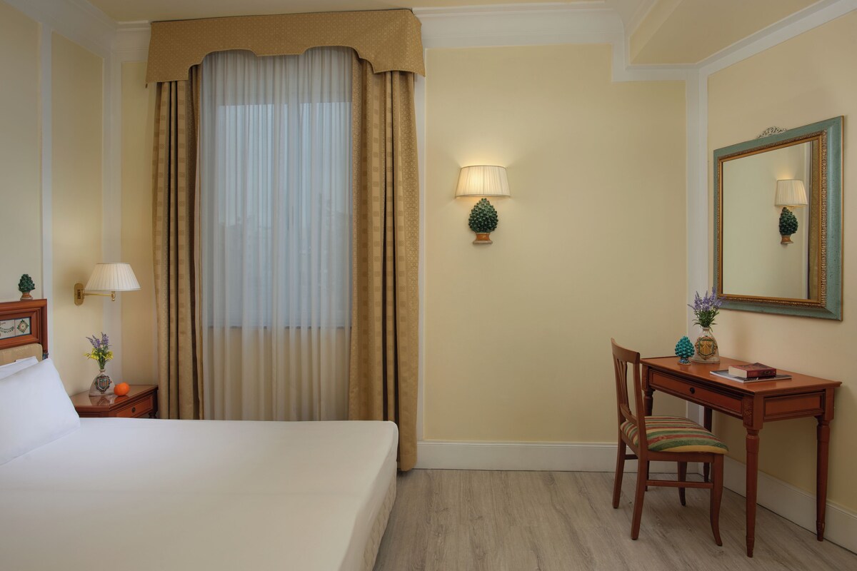 Ein Hotelzimmer mit Bett, Schreibtisch, Stuhl, Nachttisch, Lampe und Spiegel.