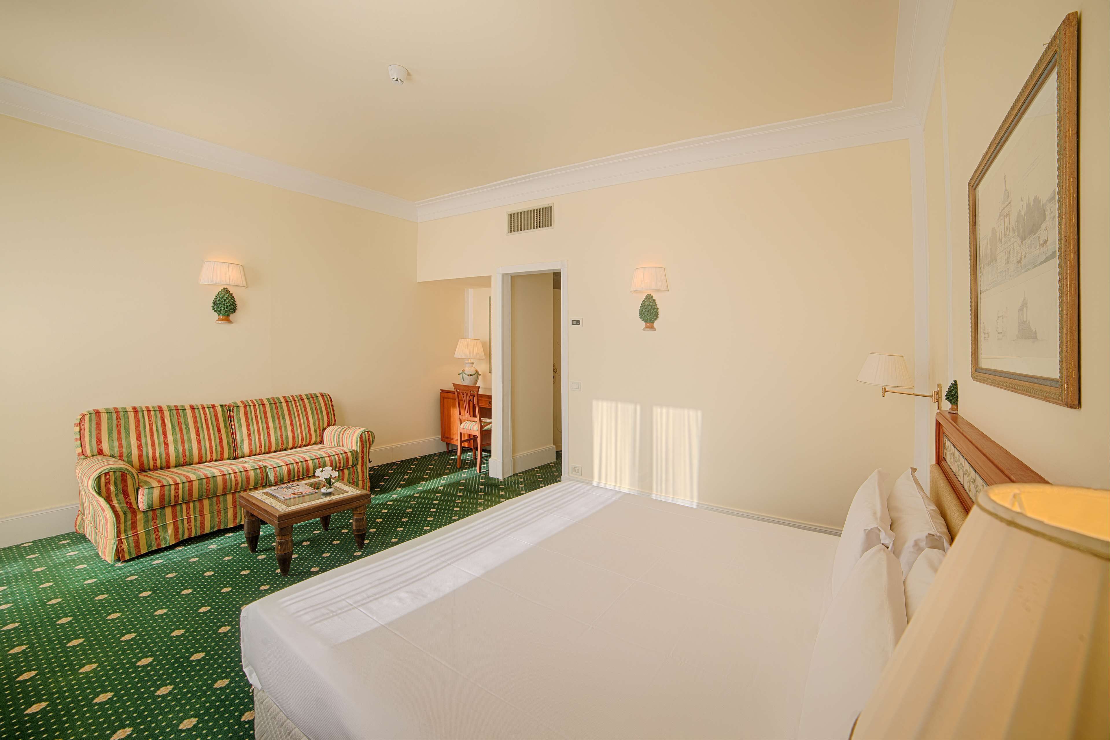 Habitación de hotel con una cama, un sofá a rayas, una mesita, dos lámparas fijadas a la pared y una puerta que da a otra habitación.
