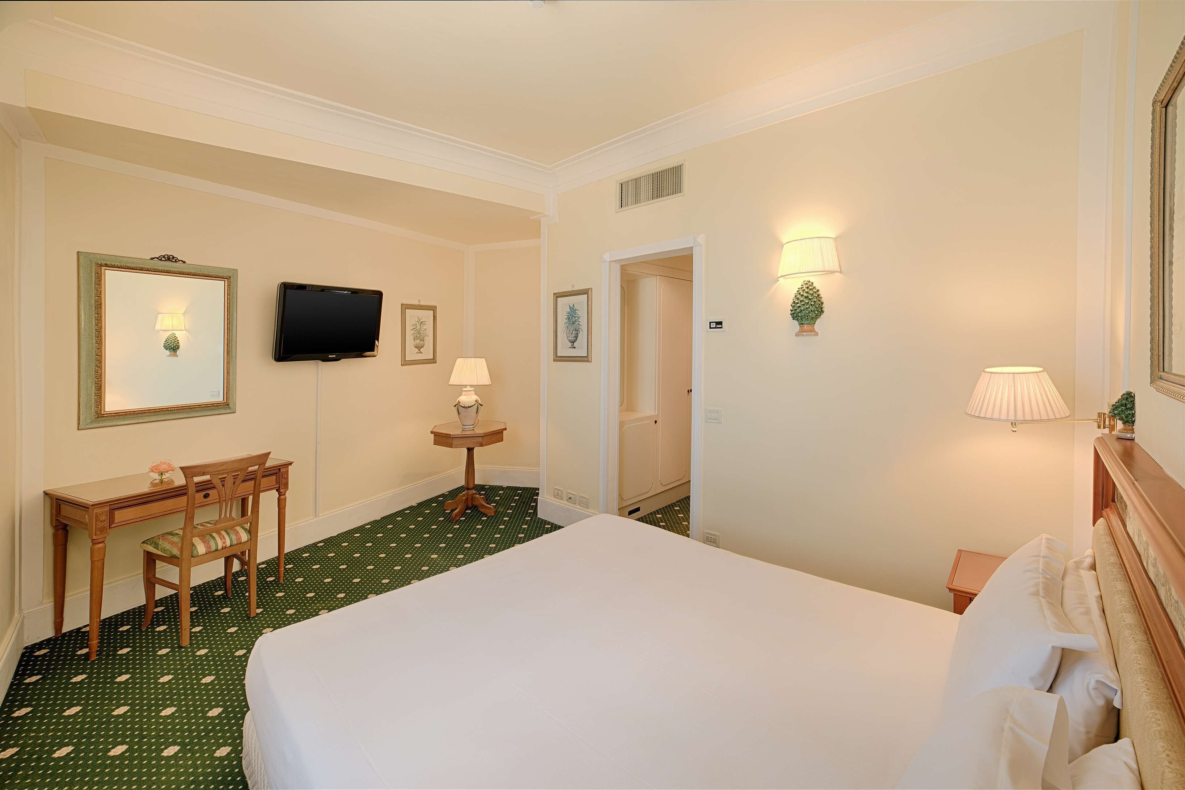 A hotel room with a bed, a desk, a chair, a television, and a small table with a lamp.