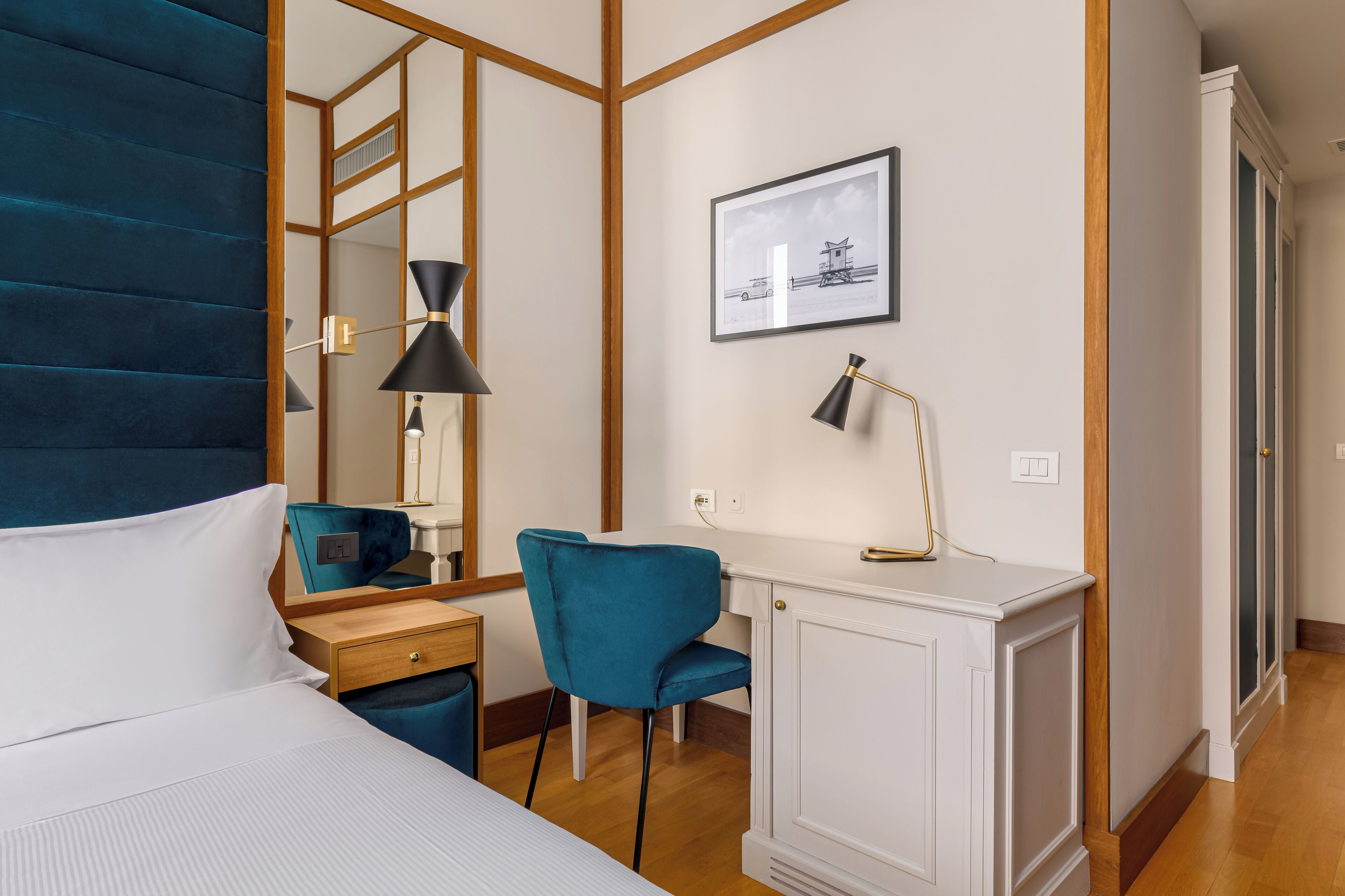 Habitación de hotel con cama, escritorio con silla, espejo, un cuadro enmarcado en la pared y armario.