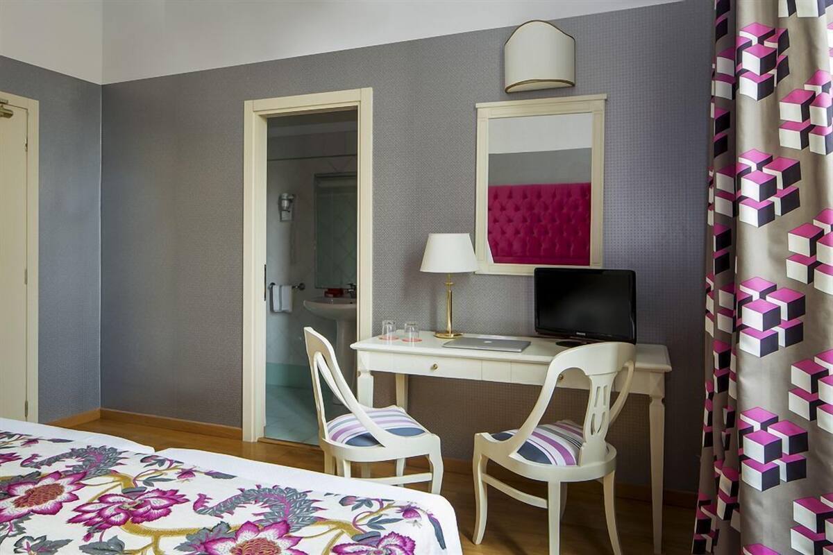 Une chambre d’hôtel avec un lit, un bureau avec un ordinateur, deux chaises, un miroir et une salle de bain visible à travers une porte ouverte.