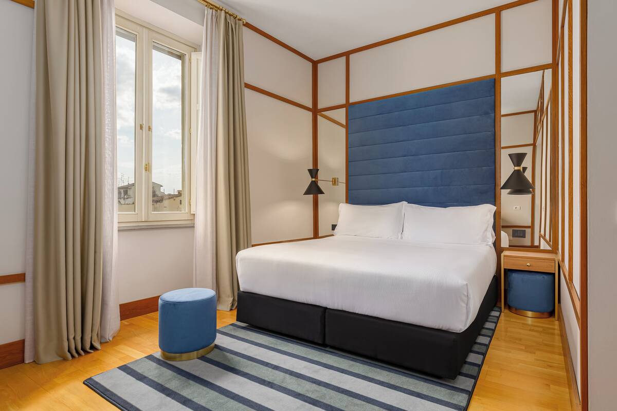 Une chambre à coucher bien rangée, avec un grand lit, une table de chevet, une lampe et une fenêtre avec des rideaux.