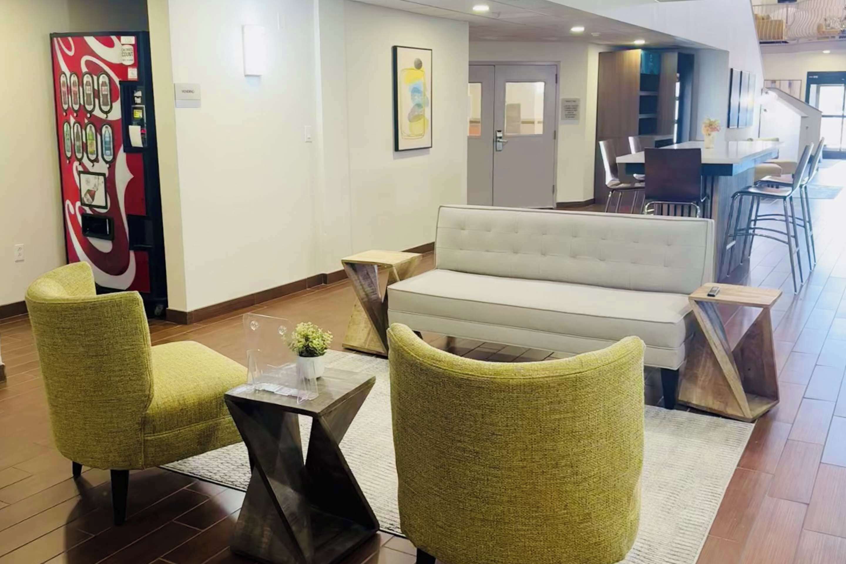 Un hall de hotel con un sofá, dos sillones, una cafetera y un comedor con sillas y una mesa.