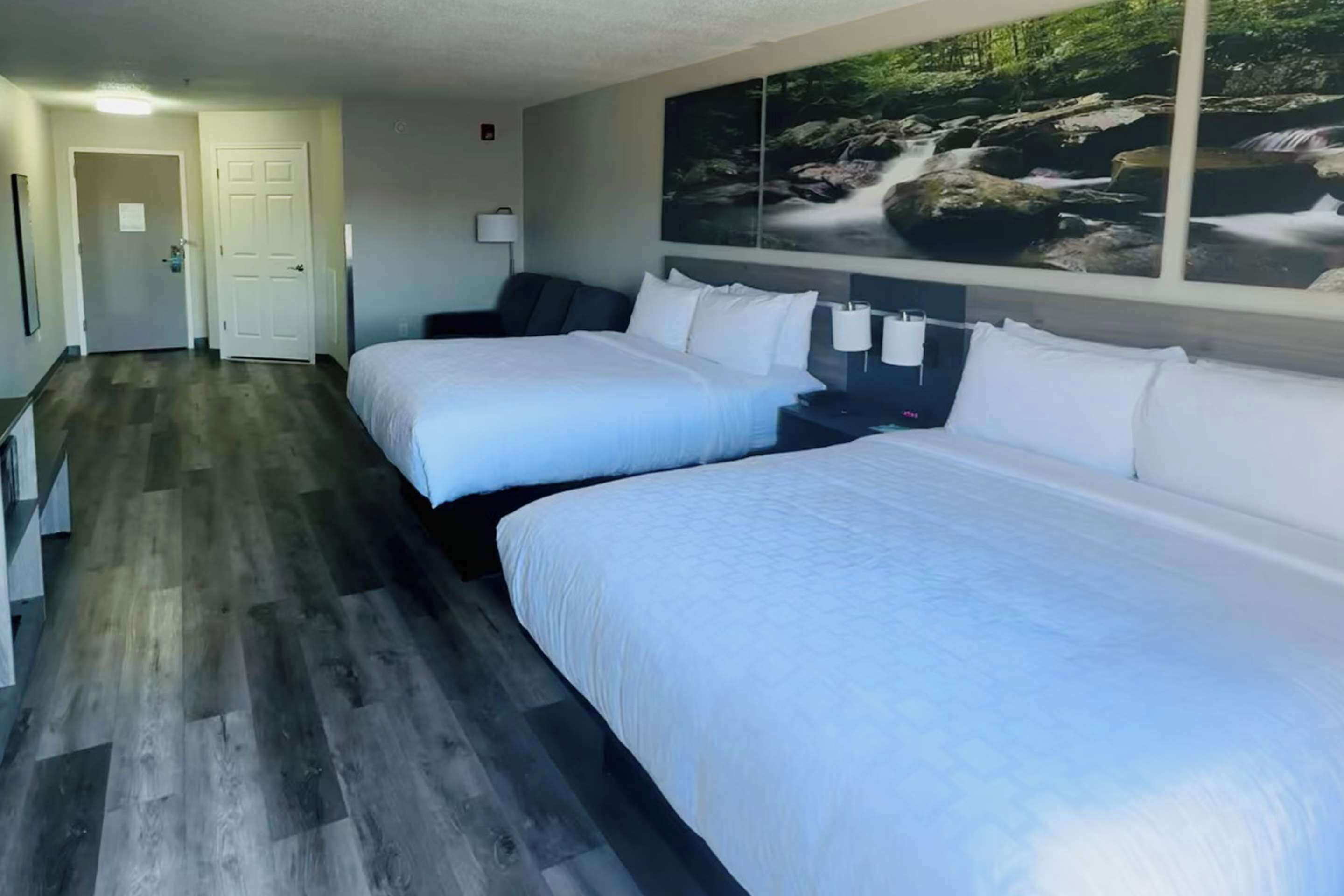 A hotel room with two beds, a wooden floor, a painting of a waterfall on the wall, and a door in the background.