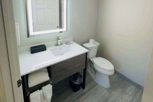 A modern bathroom with a white sink, dark wooden vanity, a mirror with a light, a toilet, and a trash bin.