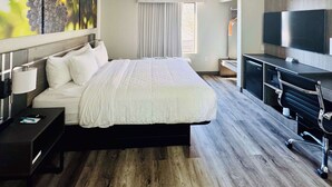 A hotel room with a large bed, a desk with a TV, and a painting of grapes on the wall. - Clarion Pointe Albemarle (Albemarle)