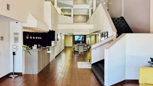 A spacious lobby with a wooden floor, a reception desk, a staircase, and a dining area with a TV screen. - Clarion Pointe Albemarle (Albemarle)