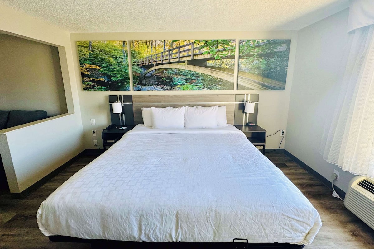 A hotel room with a large bed, a headboard with a painting of a bridge, two bedside tables with lamps, and a window with curtains.