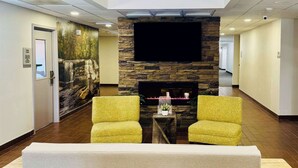 A lobby with a stone fireplace, two yellow armchairs, a sofa, and a flat-screen TV mounted above the fireplace. - Clarion Pointe Albemarle (Albemarle)
