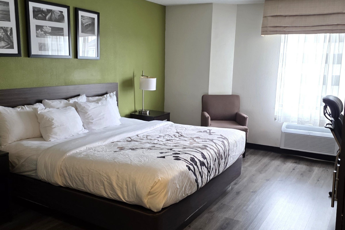 A neatly made bed with white linens, a bedside table with a lamp, a chair, and framed pictures on the wall.