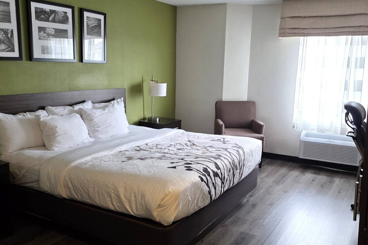 A neatly made bed with white linens, a bedside table with a lamp, a chair, and framed pictures on the wall.