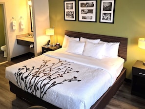 Une chambre d’hôtel comprenant un lit, des tables de chevet, une petite table, une lampe, une salle de bain avec un lavabo et un porte-serviettes, ainsi que des tableaux encadrés accrochés au mur.