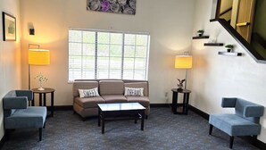 Een woonkamer met een bank, twee fauteuils, een salontafel, een raam met zonwering, een schilderij en twee vloerlampen.
