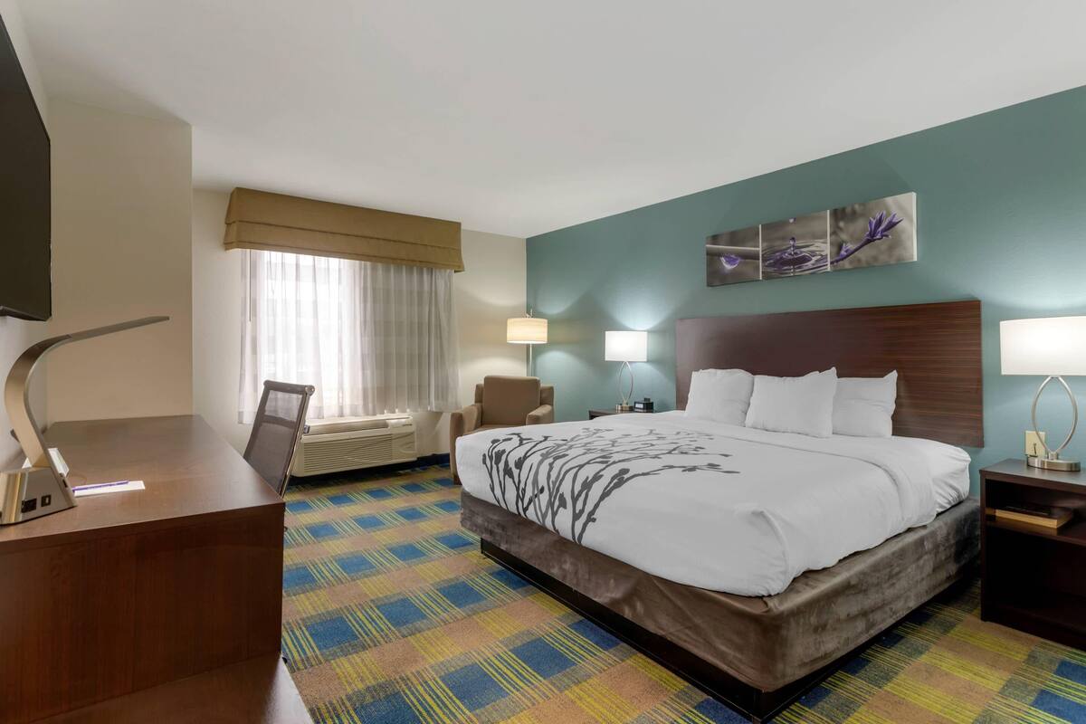 A hotel room with a large bed, a desk, a chair, a window with curtains, and wall-mounted lamps.
