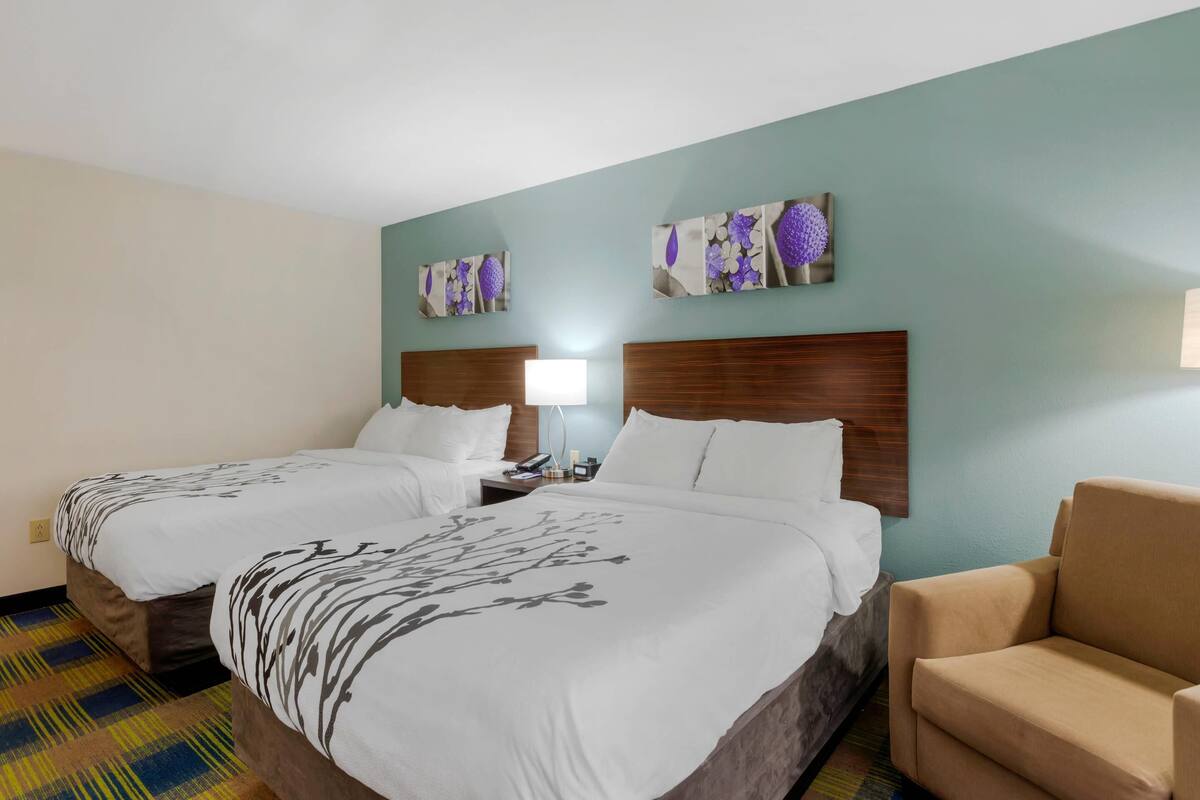 A hotel room with two beds, a wooden headboard, a nightstand, a lamp, and a chair.