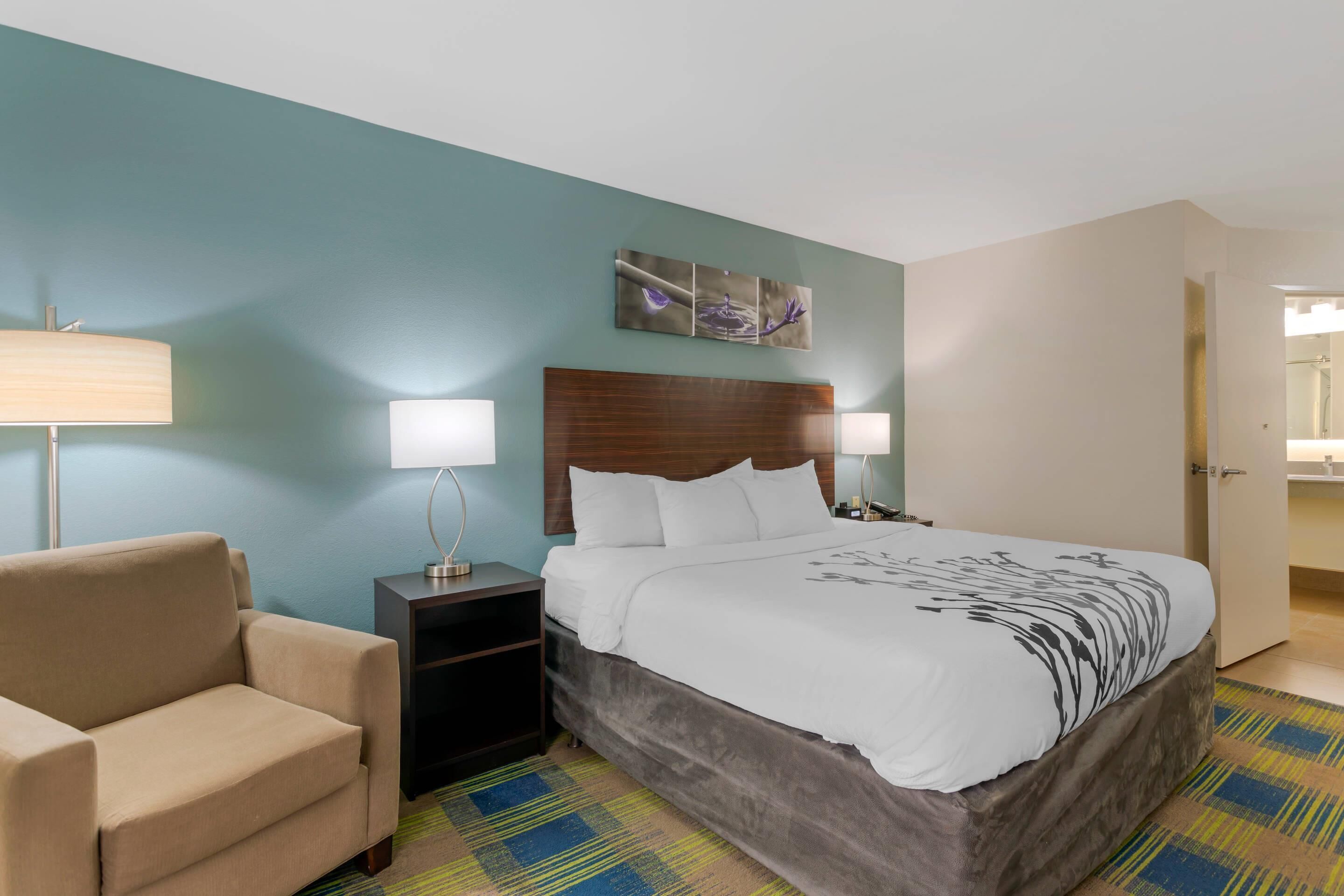 A hotel room with a bed, a nightstand, a lamp, a chair, and a wall with a painting.