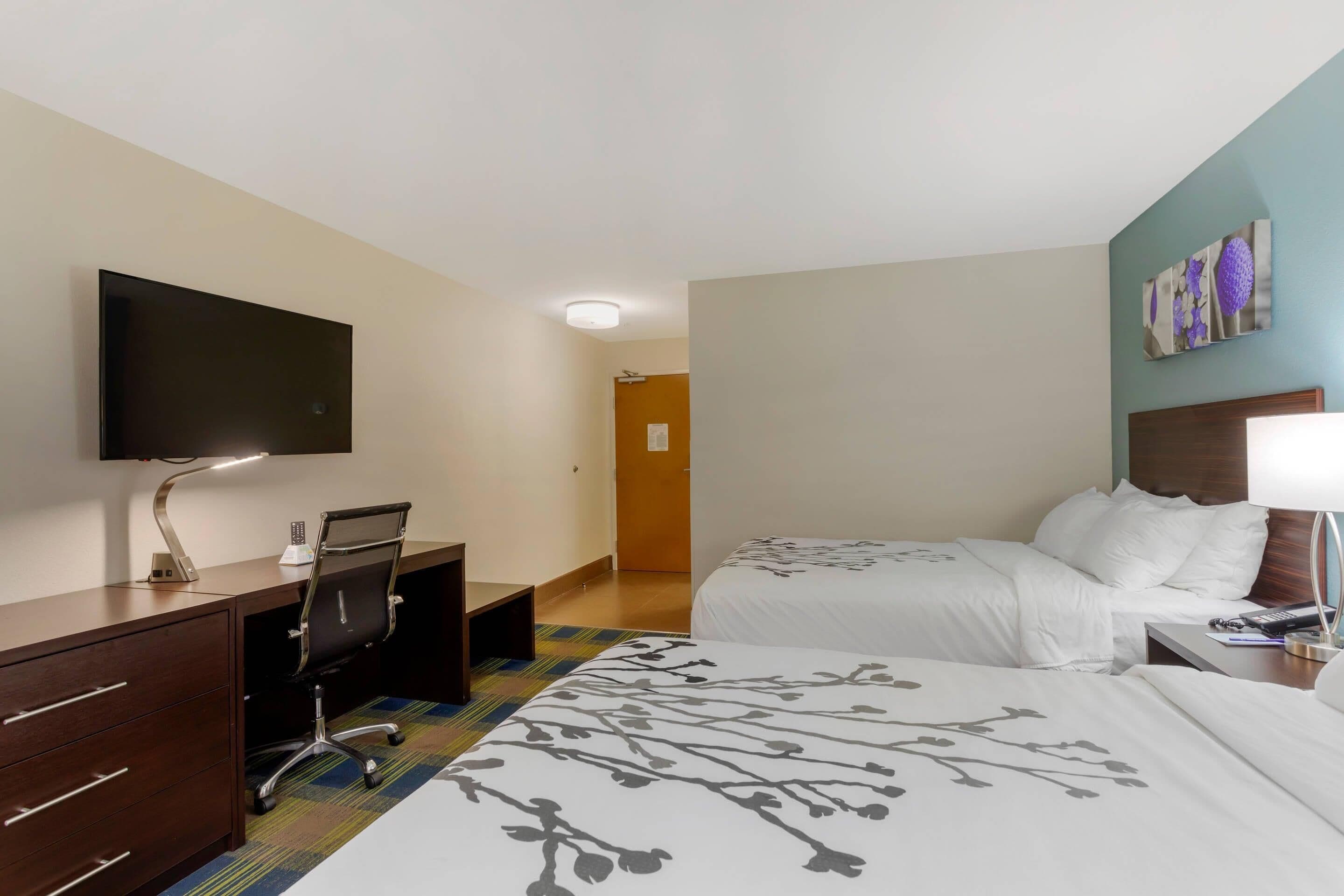 A hotel room with a desk, two beds, a TV, and a painting on the wall.