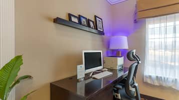 A hotel room with a desk, computer, chair, lamp, and framed pictures on the wall.