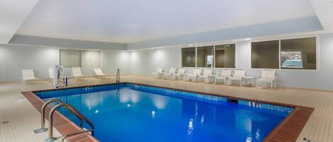 A rectangular indoor swimming pool with a tiled floor and a metal ladder. There are white lounge chairs arranged around the pool, and a window with frosted glass panels on the wall.