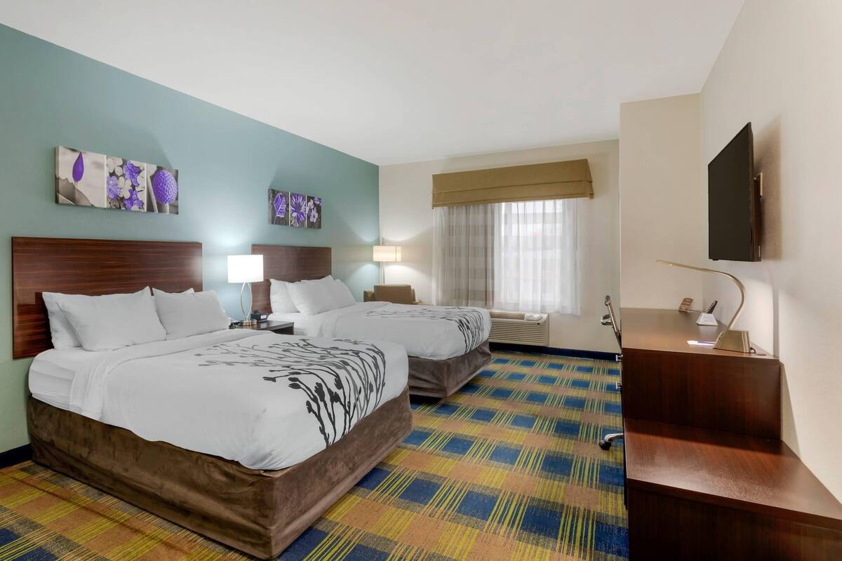 A hotel room with two beds, a desk, a flat-screen TV, and artwork on the walls.