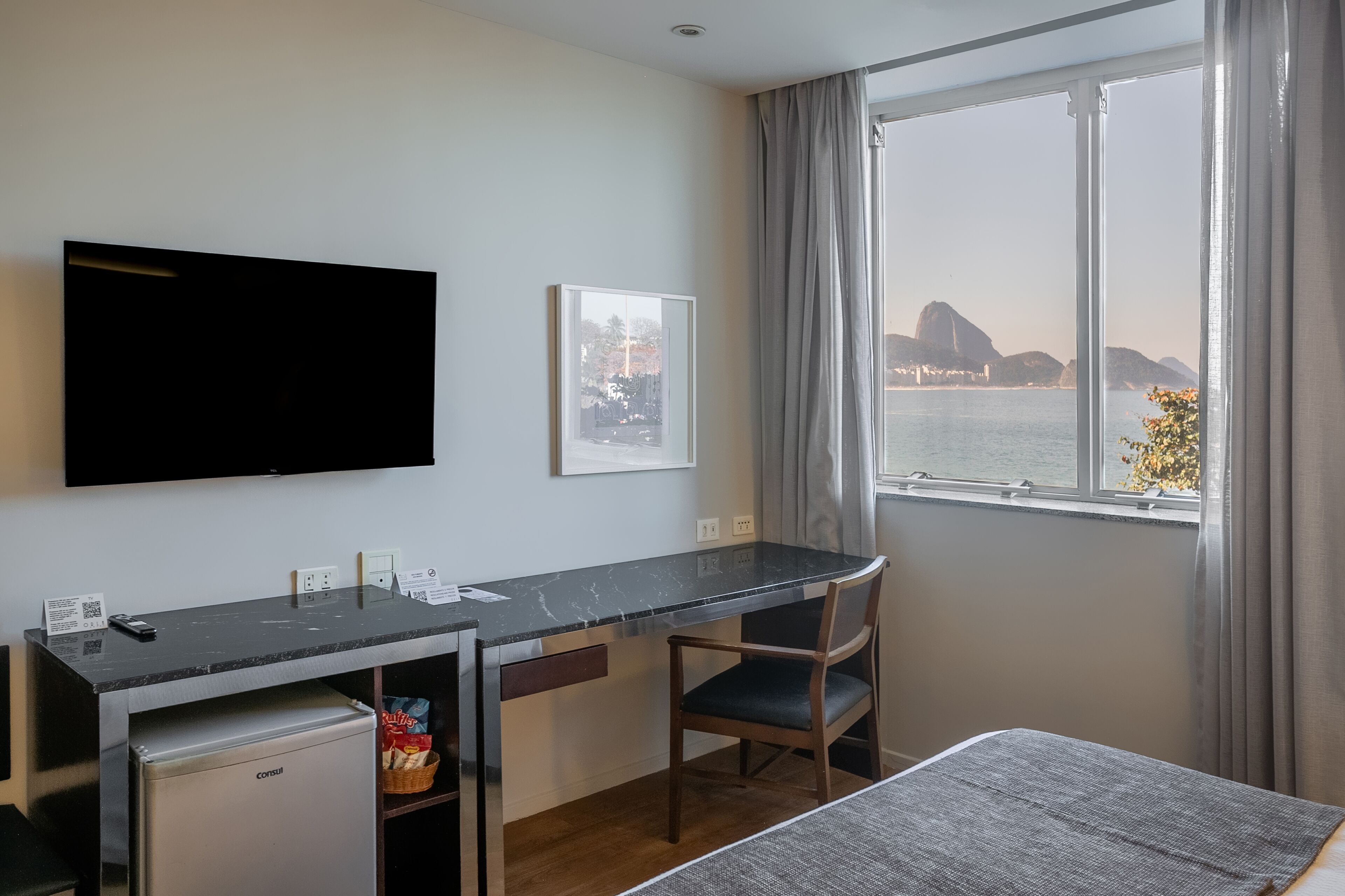 Habitación de hotel con escritorio, silla y televisor. Se ve una ventana con vistas a un cuerpo de agua y una montaña.