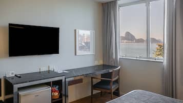 Habitación de hotel con escritorio, silla y televisor. Se ve una ventana con vistas a un cuerpo de agua y una montaña.