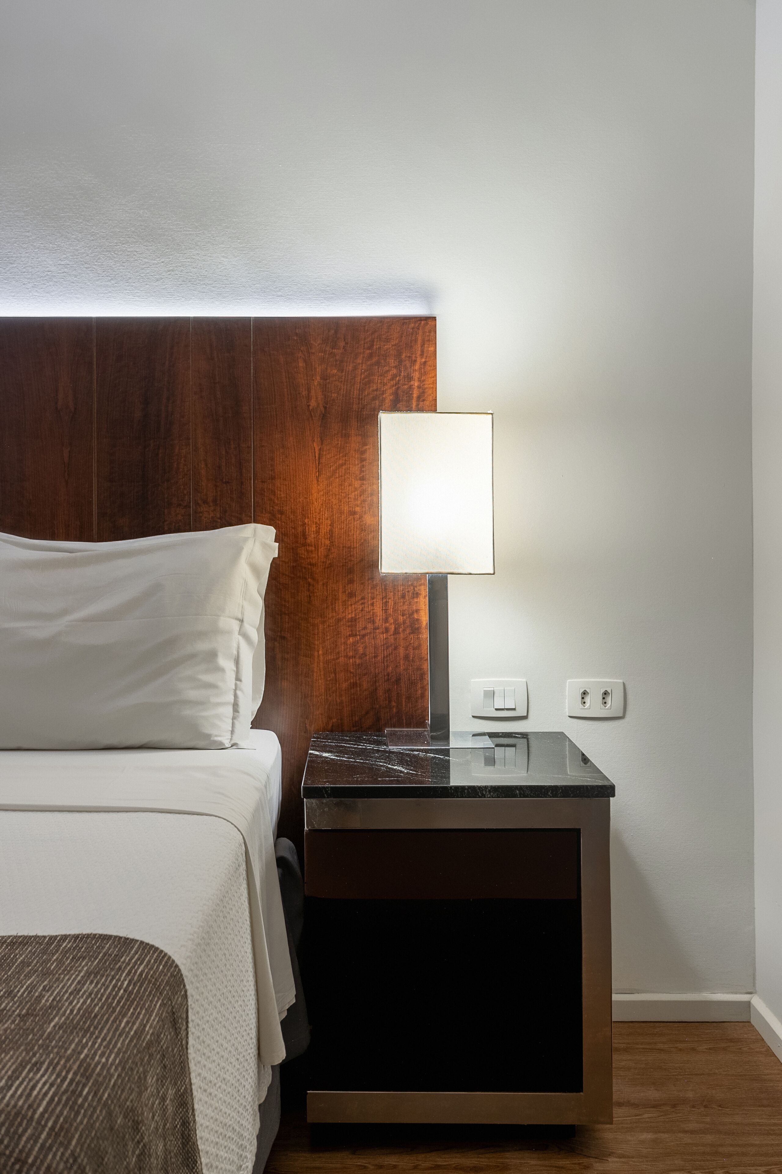 A hotel room with a wooden headboard, a bedside table with a lamp, and a power outlet.