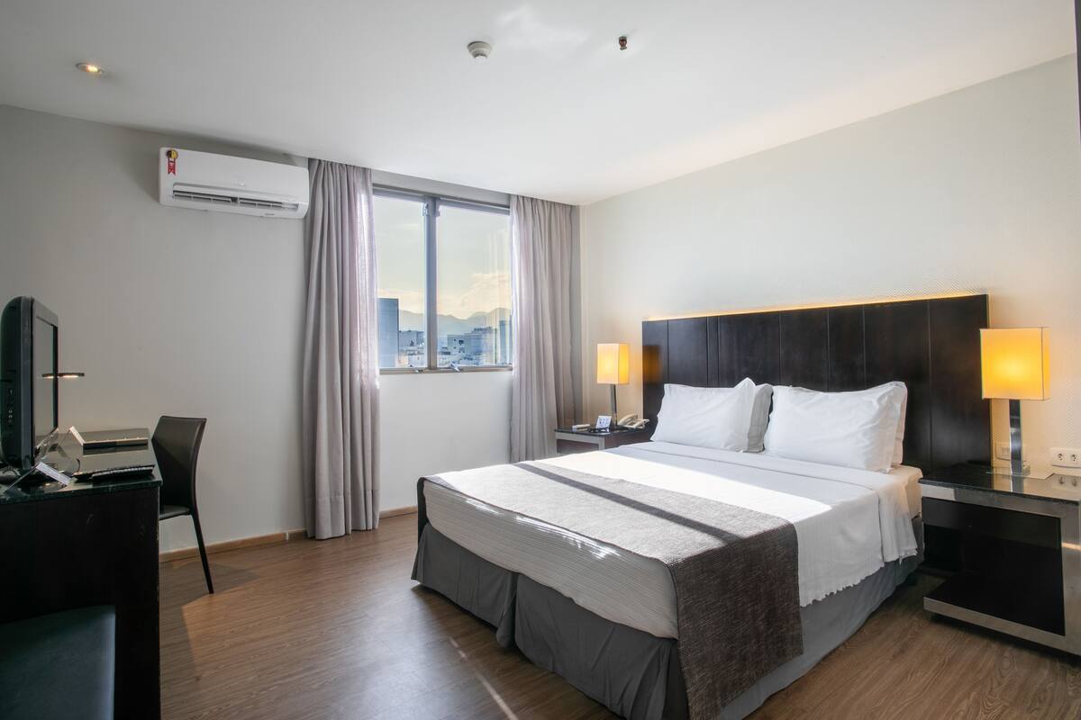 Een hotelkamer met een groot bed, een bureau met een computer, airconditioning, een raam met gordijnen en uitzicht op de stad.