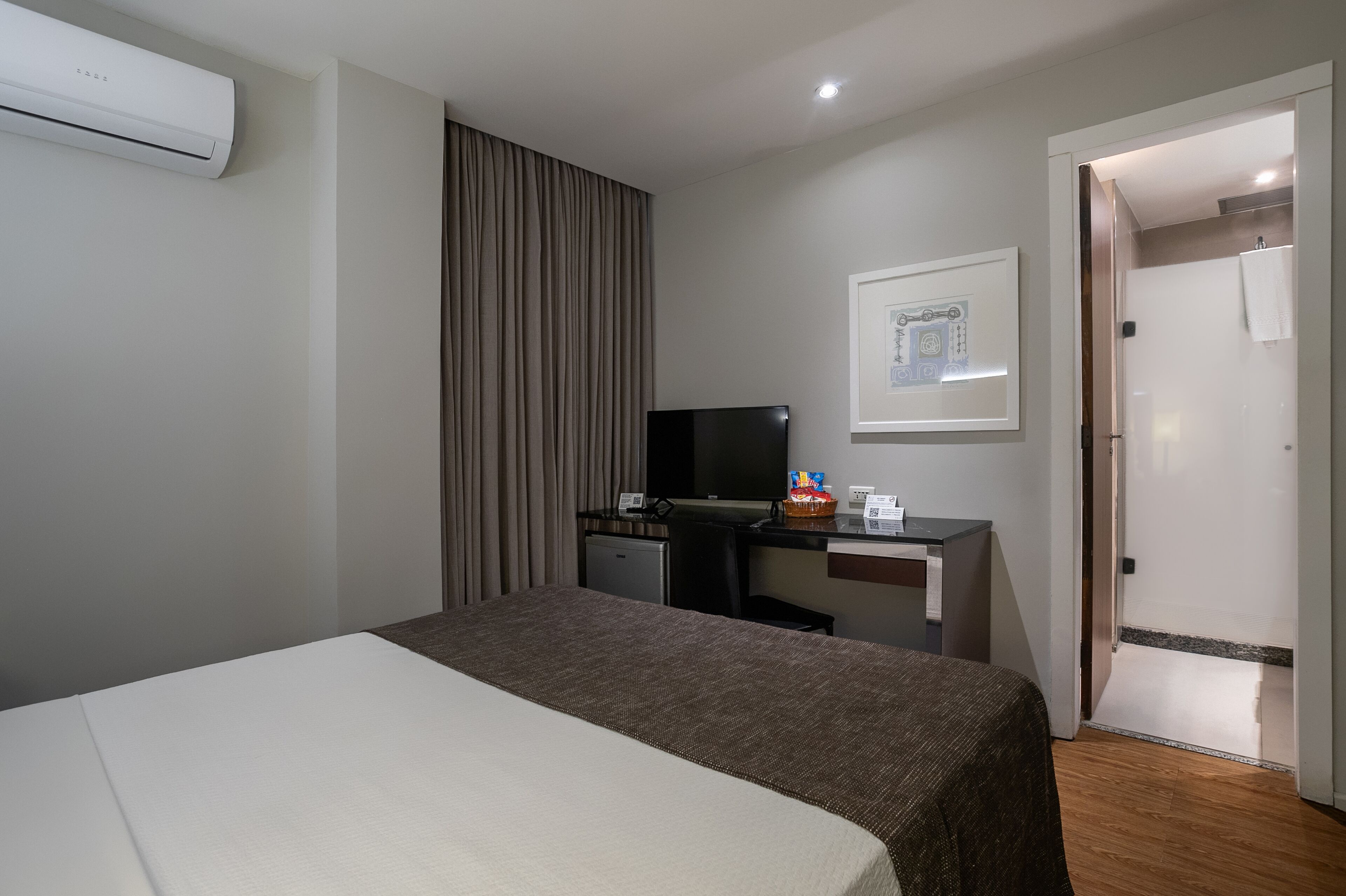 Quarto de hotel com cama, escrivaninha com computador, televisão e chuveiro.