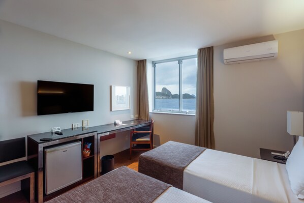 A hotel room with a bed, a desk, a television, a window with a view of a mountain, and an air conditioning unit. - Orla Copacabana Hotel (Rio de Janeiro)