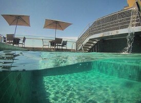 Piscina externa com água cristalina, dois guarda-sóis e uma escadaria que leva a uma área coberta.