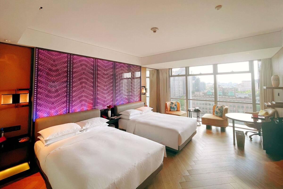 Habitación de hotel con dos camas, un escritorio, una silla, un ventanal grande y un panel decorativo en la pared.