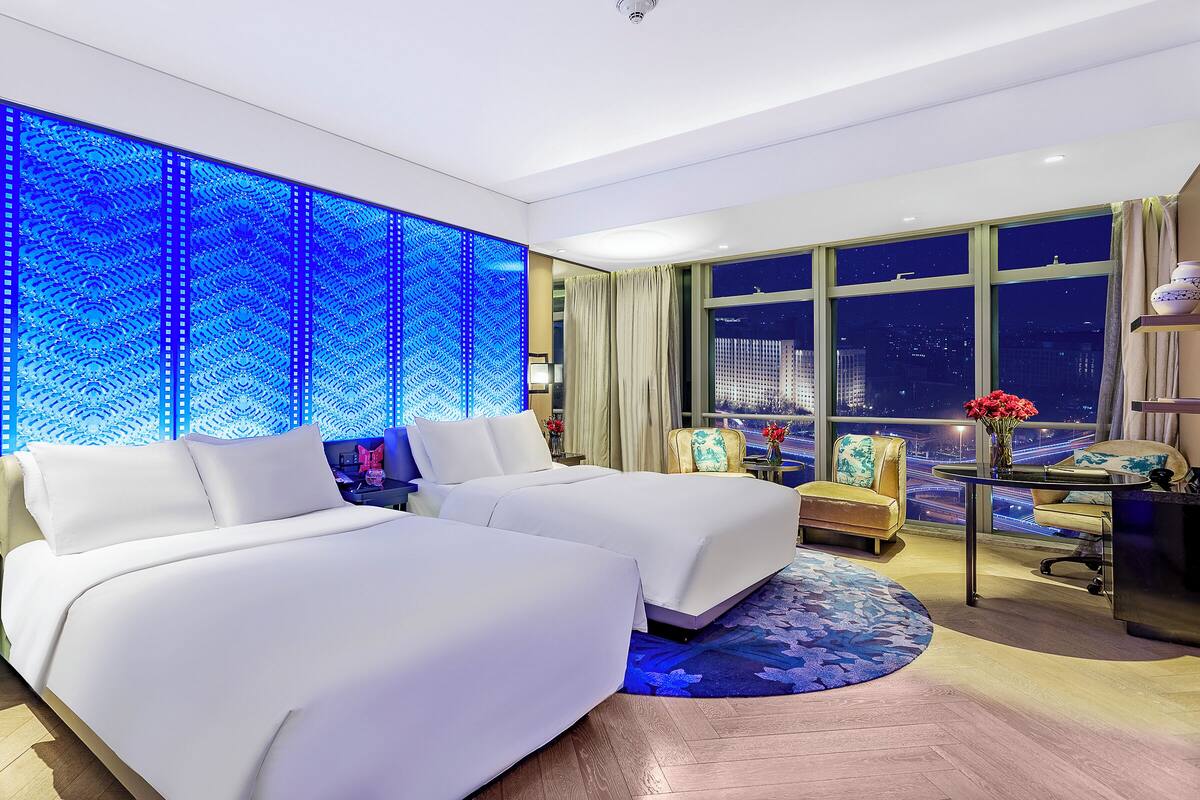 Una habitación de hotel moderna con dos camas, un ventanal con vistas a la ciudad y un panel de luz azul en la pared.