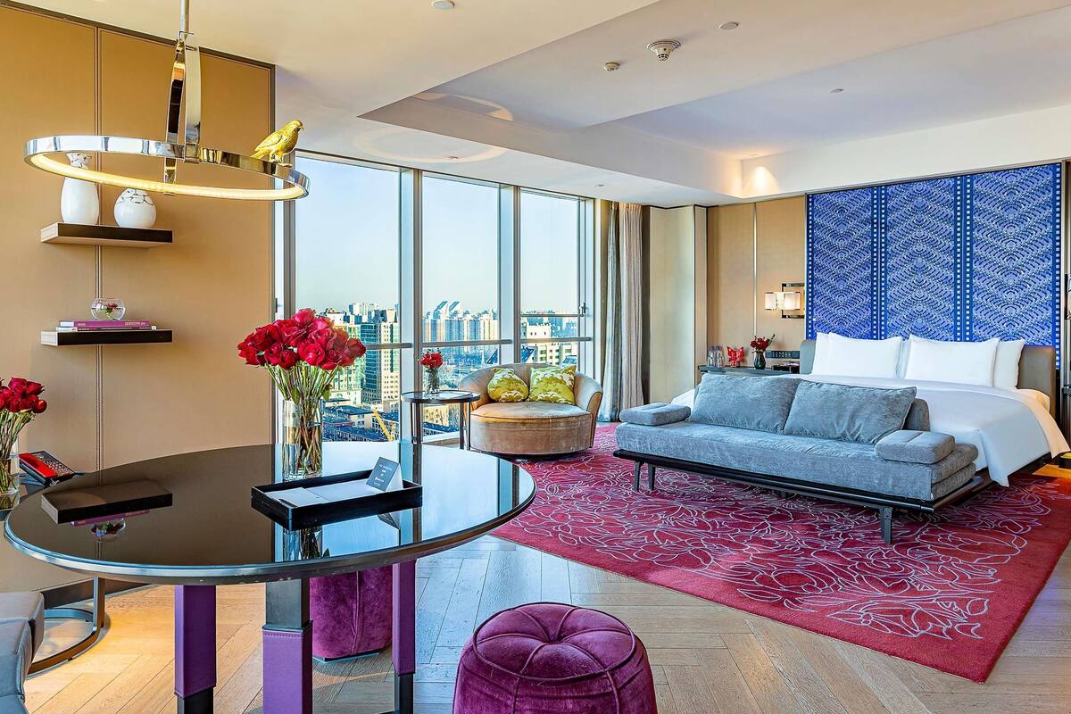 Una habitación de hotel moderna con un ventanal grande, una mesa redonda de cristal, un sofá, un tapete estampado rojo y un tapiz decorativo en la pared.