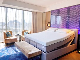 Una habitación de hotel moderna con una cama grande, una zona de estar con una silla y una mesita, y vistas de la ciudad a través de amplios ventanales.