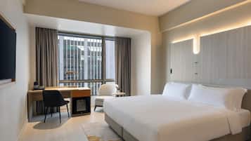 Quarto de hotel moderno com uma cama grande, uma mesa com cadeira, uma mesinha e uma janela com cortinas.