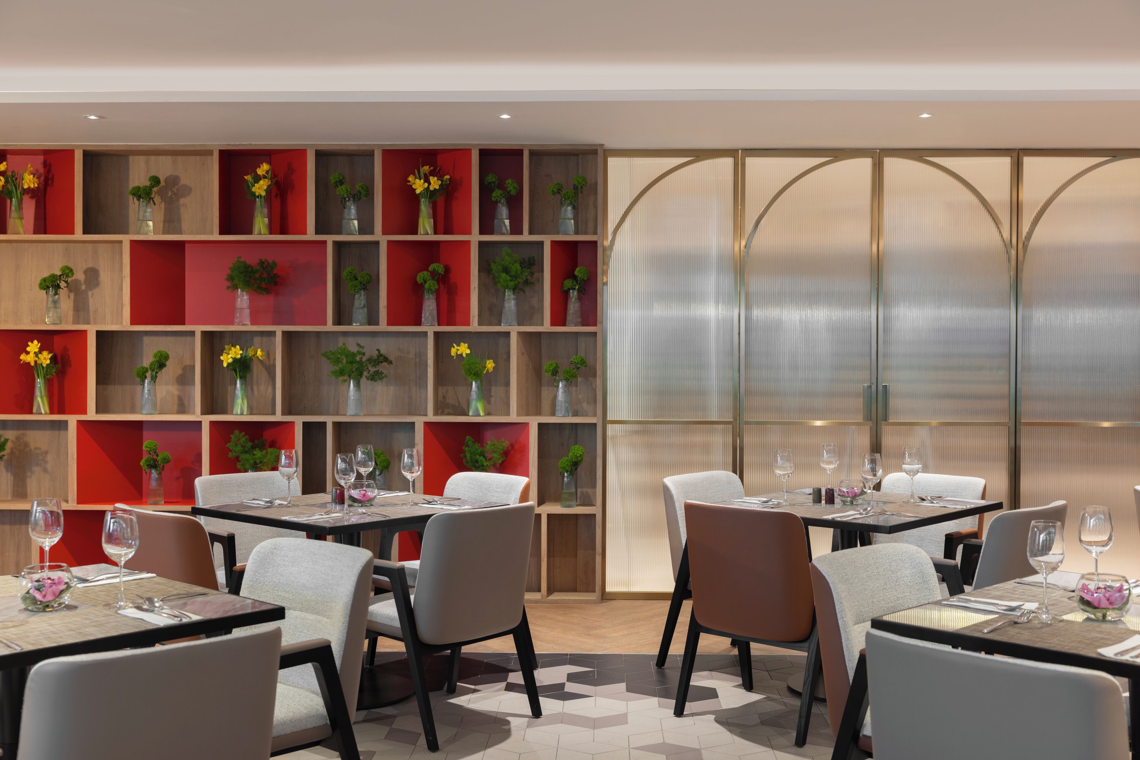 Un restaurante moderno con varias mesas y sillas, estantes rojos y de madera con flores, y un muro divisorio de vidrio.