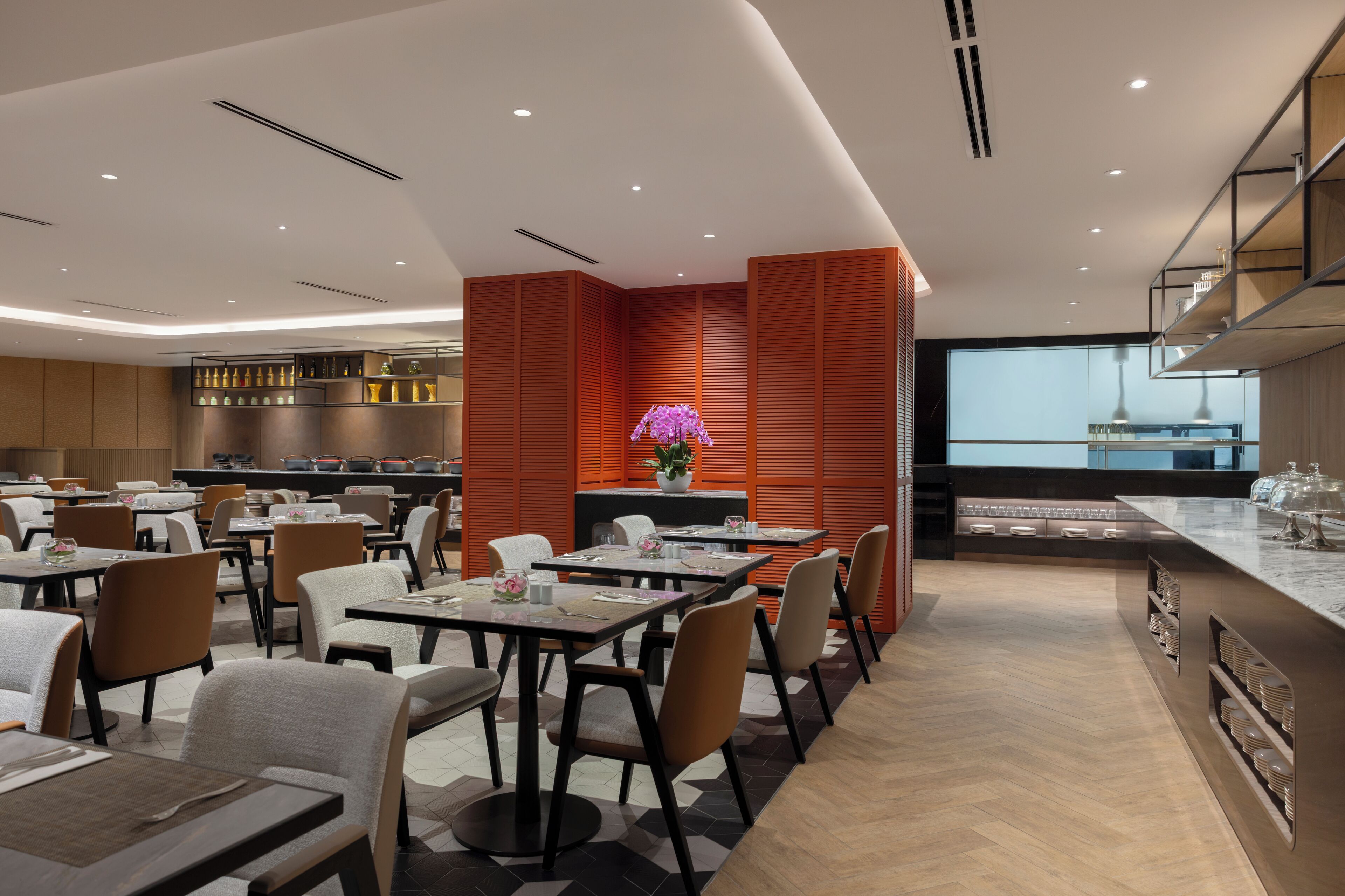 A modern restaurant with multiple dining tables and chairs, a central floral arrangement, and a kitchen area visible in the background.