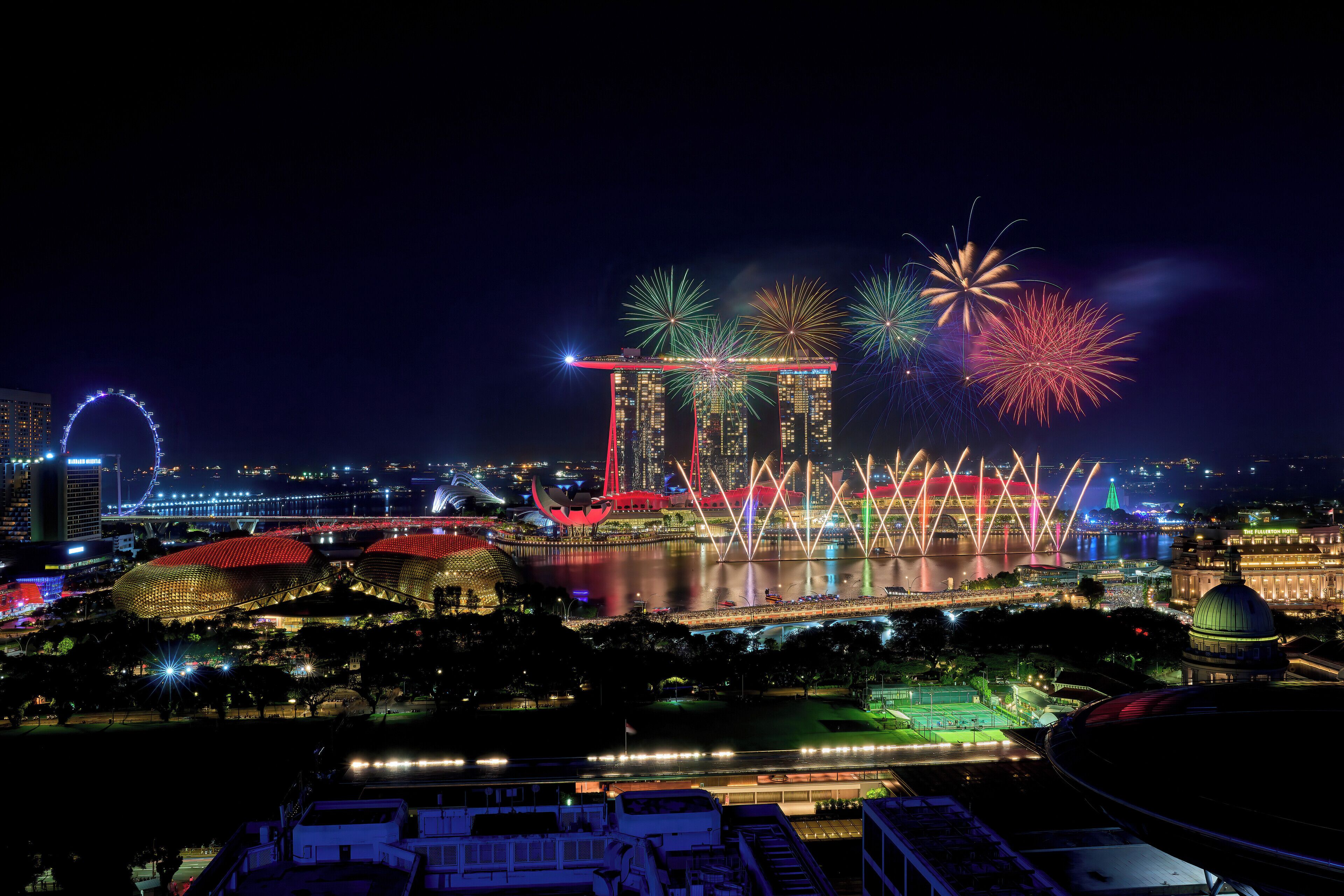 A nighttime cityscape with illuminated buildings, a prominent bridge, and fireworks.