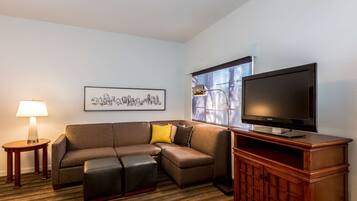 Habitación de hotel con sofá modular, televisor sobre un mueble de madera, lámpara y un cuadro con una vista urbana enmarcada en la pared.