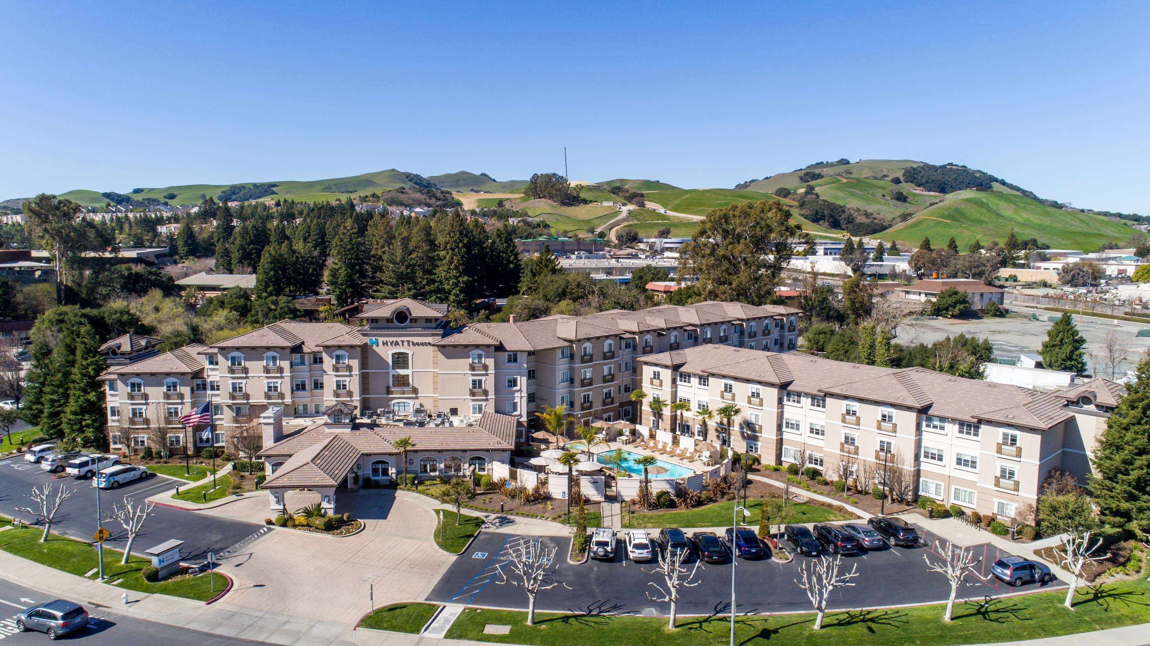 A multi-story hotel with a swimming pool, parking lot, and a view of surrounding hills.